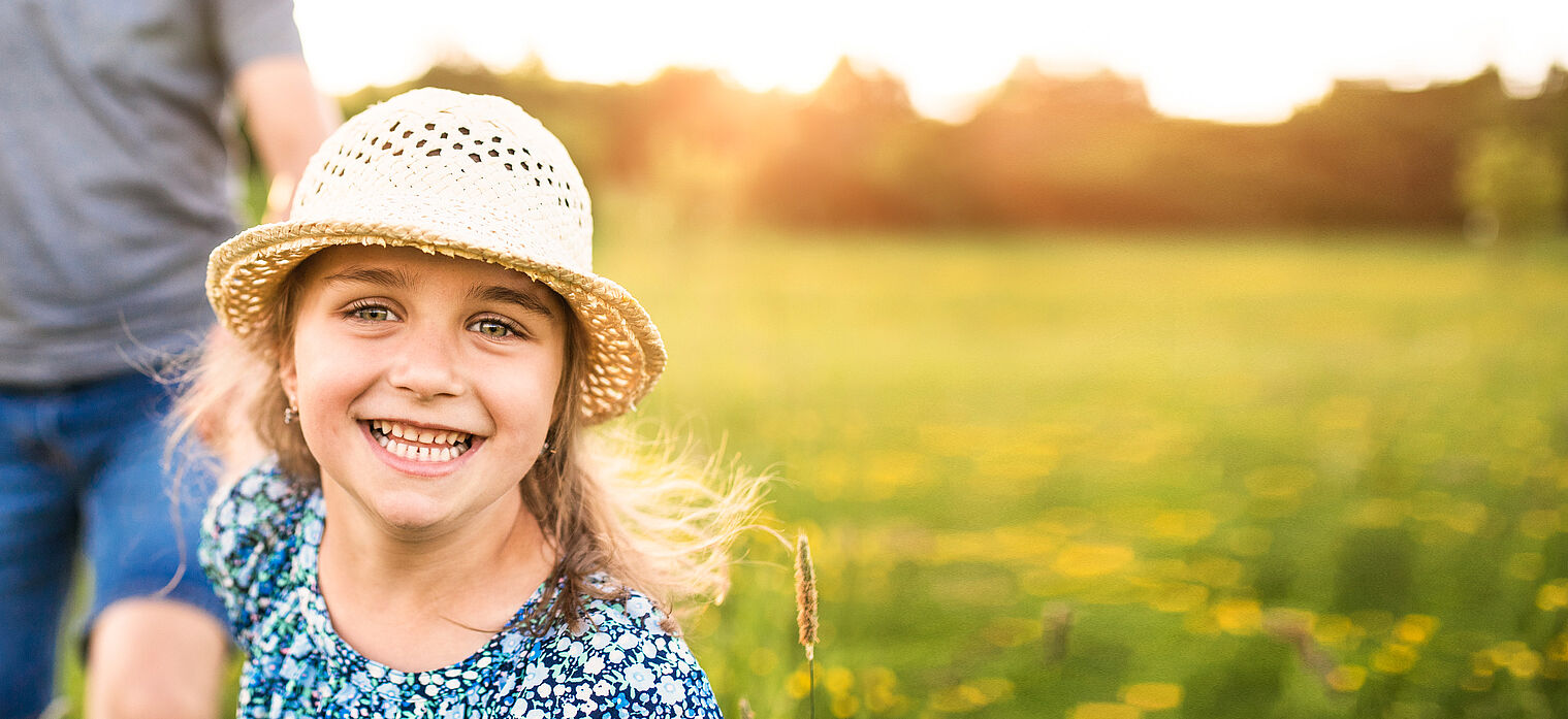 Smiling young girl wearing a straw hat and a blue floral dress, running in a sunny field with yellow flowers.