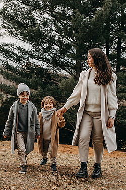 Mother and two children, all wearing coats and scarves, walking hand in hand through a forested area on a cloudy day.