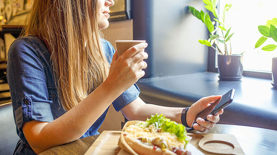 Young woman sitting at a café table with a smartphone in one hand and a cup of coffee in the other, next to a plate of healthy food.