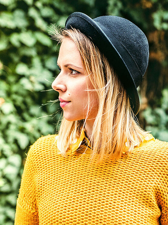 A young woman with blonde hair is wearing a black hat and a yellow sweater. She is standing in front of a background of green foliage, looking to the side