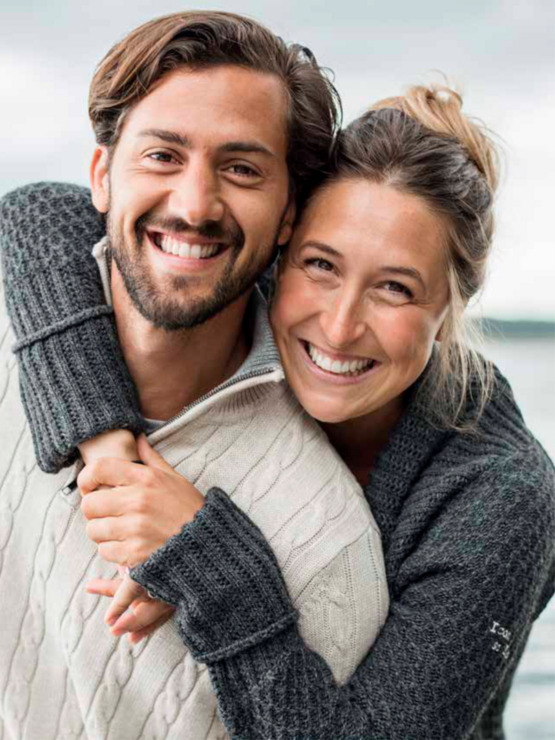 Happy couple embracing and smiling at the camera, dressed warmly in casual outdoor clothing.